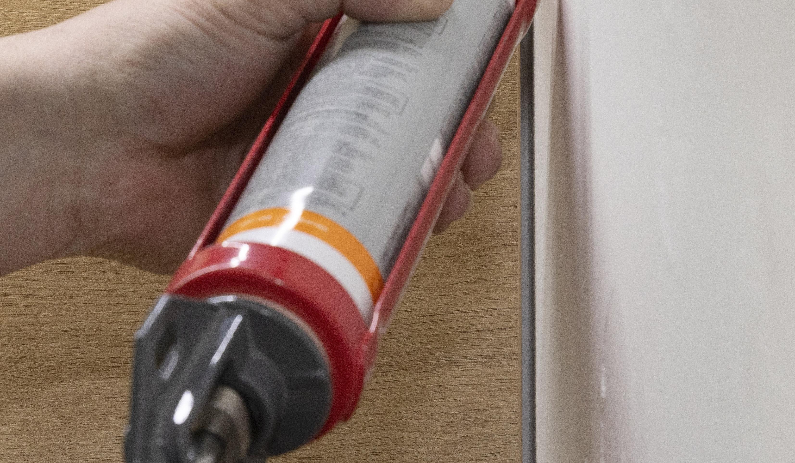 Hand trägt mit Kartuschenpistole Dichtmasse auf Fuge zwischen Wand und Holz auf.